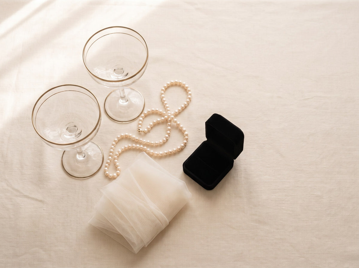 Two coupe champagne glasses, a strand of pearls, ivory tulle and a black ring box on cream linen.