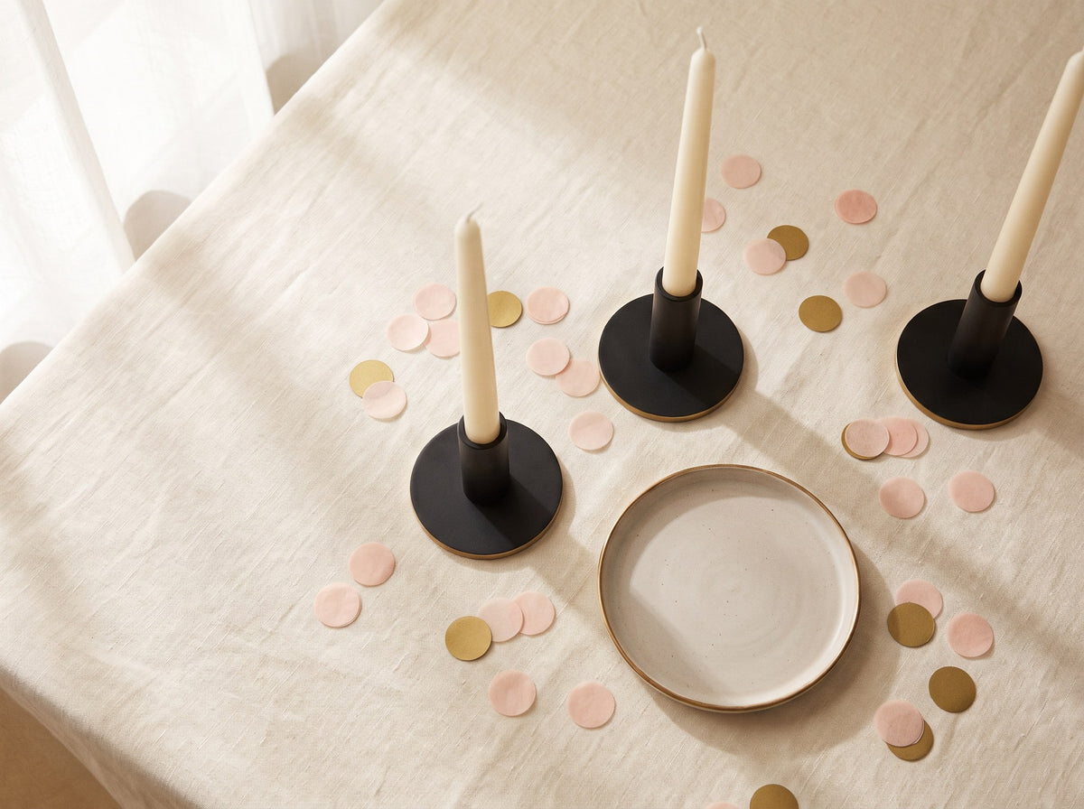 Cream taper candles in black holders with blush and gold confetti scattered on cream linen.