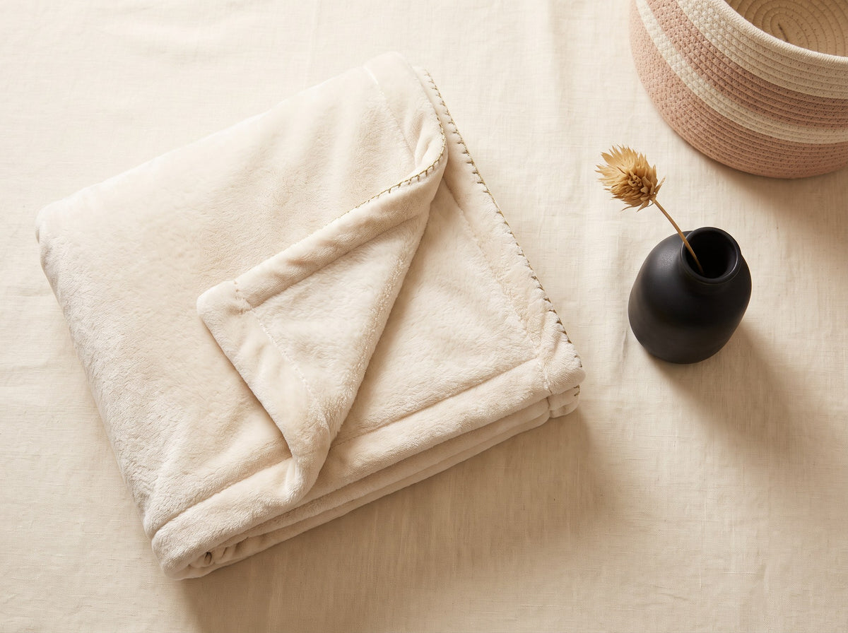A folded cream plush throw blanket beside a small black ceramic vase and a woven basket.