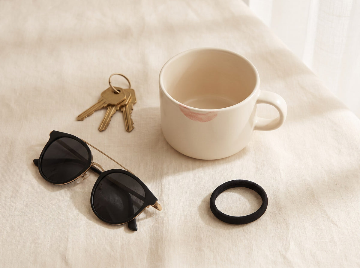 A cream coffee cup with a lipstick mark, black sunglasses, keys and a hair tie on cream linen.
