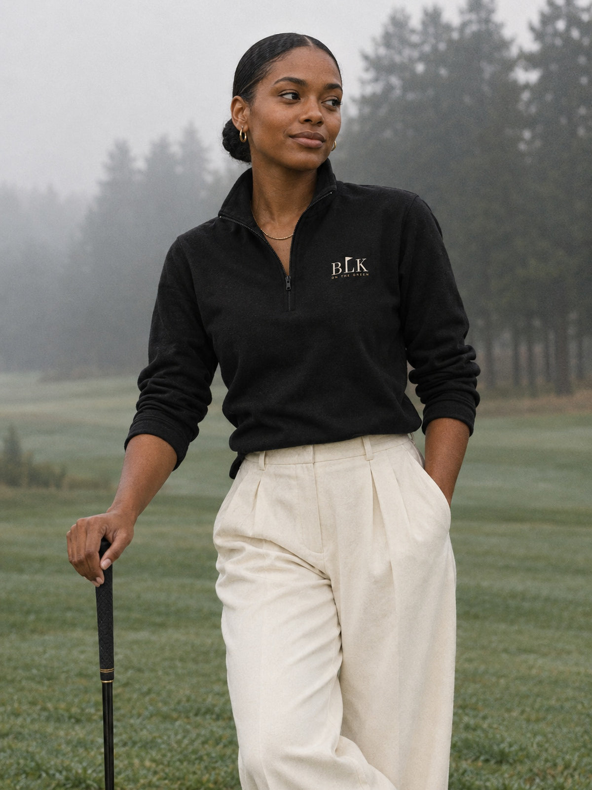 Young Black woman in the black BLK on the Green Fairway Quarter Zip and ivory trousers, leaning on a club on a misty fairway