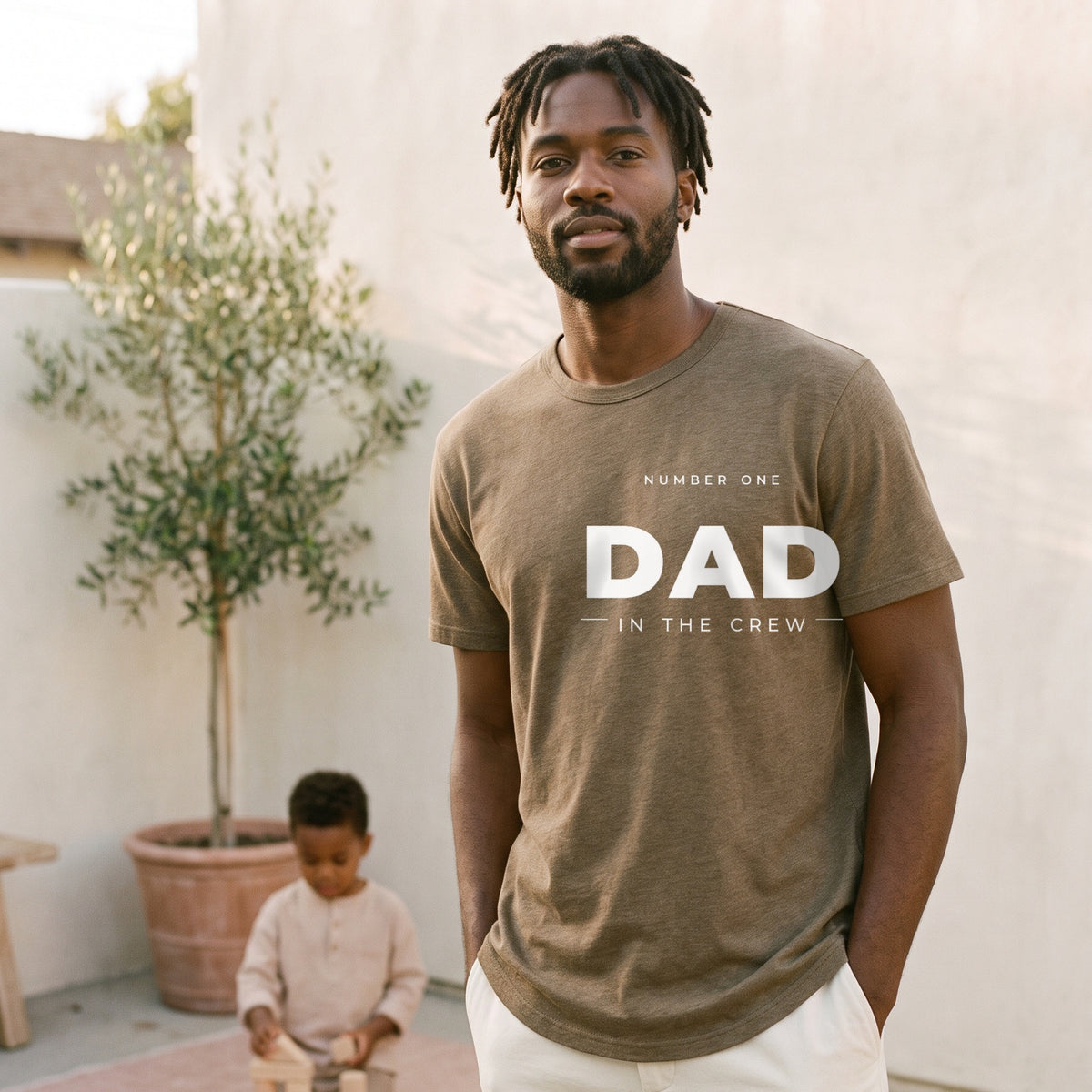 Man wearing the #1 Dad in the Crew tee in heather olive on a patio at golden hour, his son playing behind him