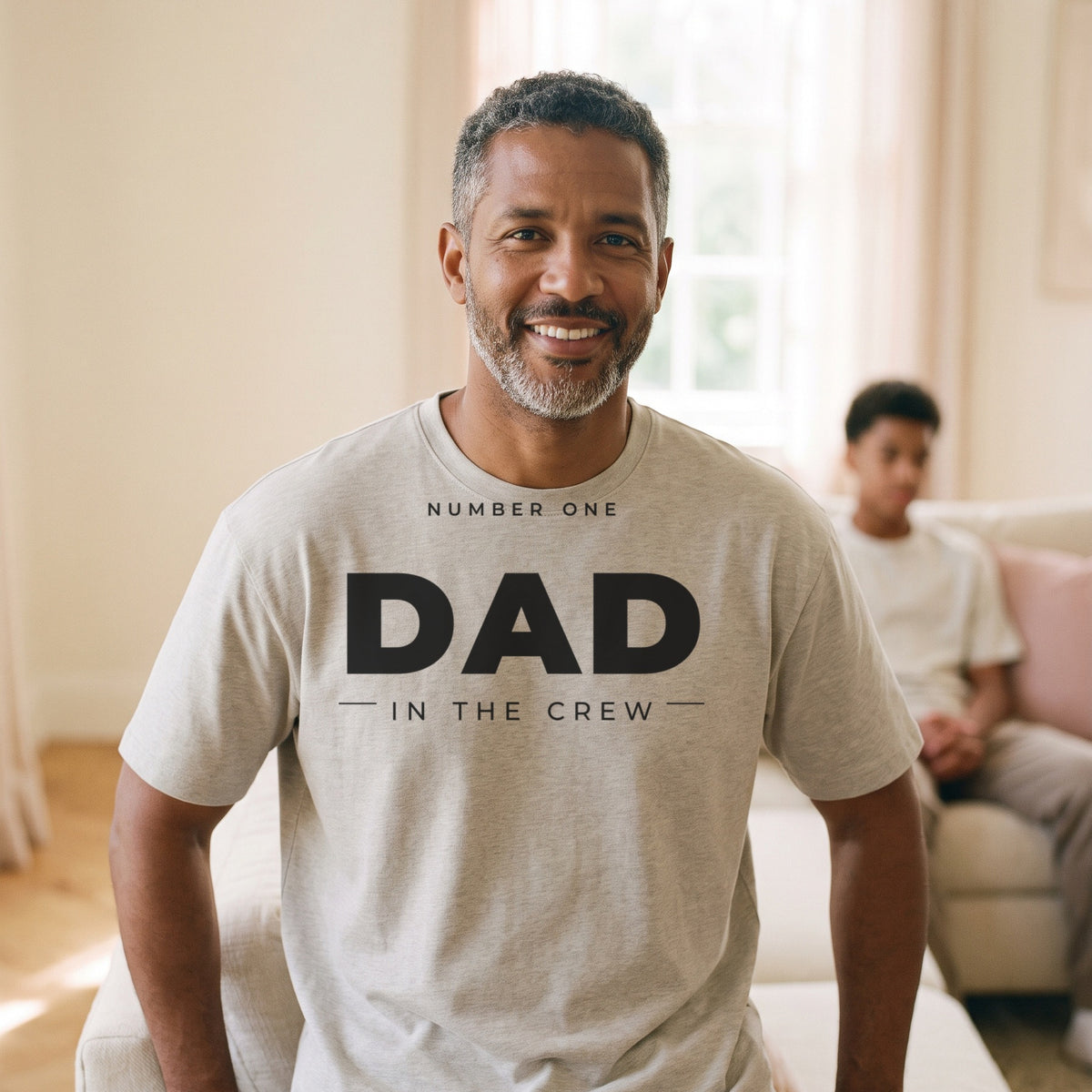 Man wearing the #1 Dad in the Crew tee in heather stone in a sunlit living room with his son
