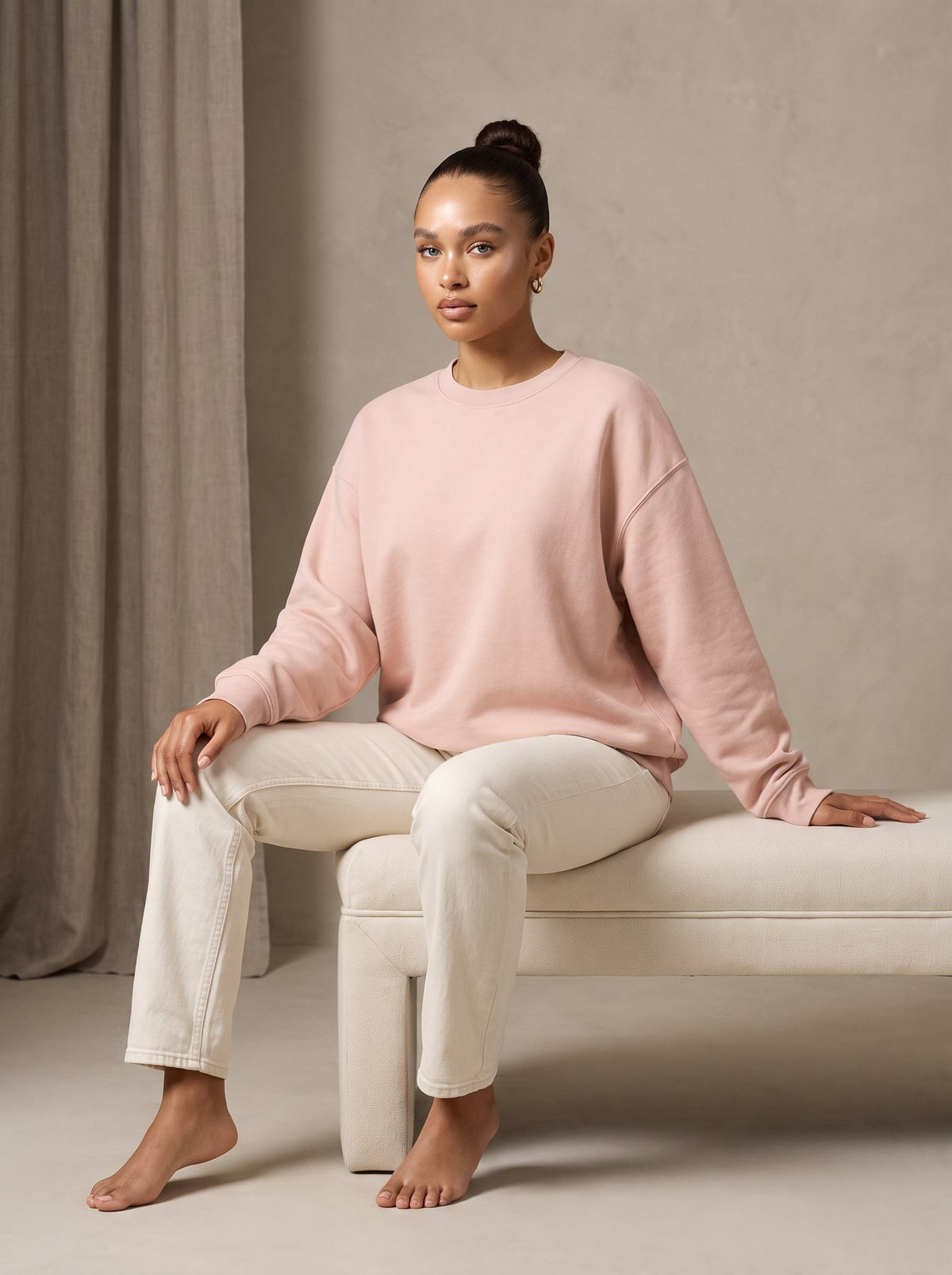 A woman in a blush crewneck seated on a cream bench in a studio