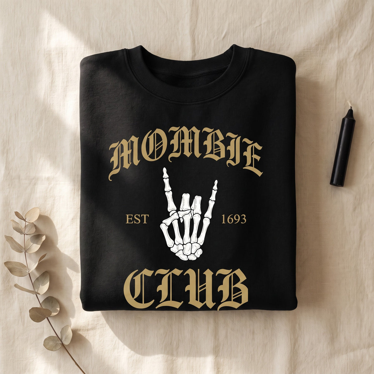 Folded black Mombie Club crewneck sweatshirt on warm cream linen, antique gold blackletter lettering with a bone white skeleton hand